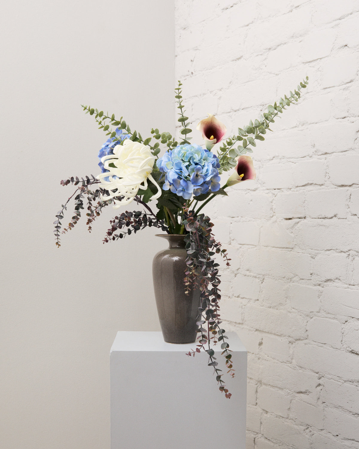 Blue Hour – bukett med konstgjorda blå hortensior, vita kallor, vit krysantemum och eukalyptus i en grå vas.
