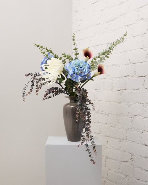 Blue Hour – bukett med konstgjorda blå hortensior, vita kallor, vit krysantemum och eukalyptus i en grå vas.