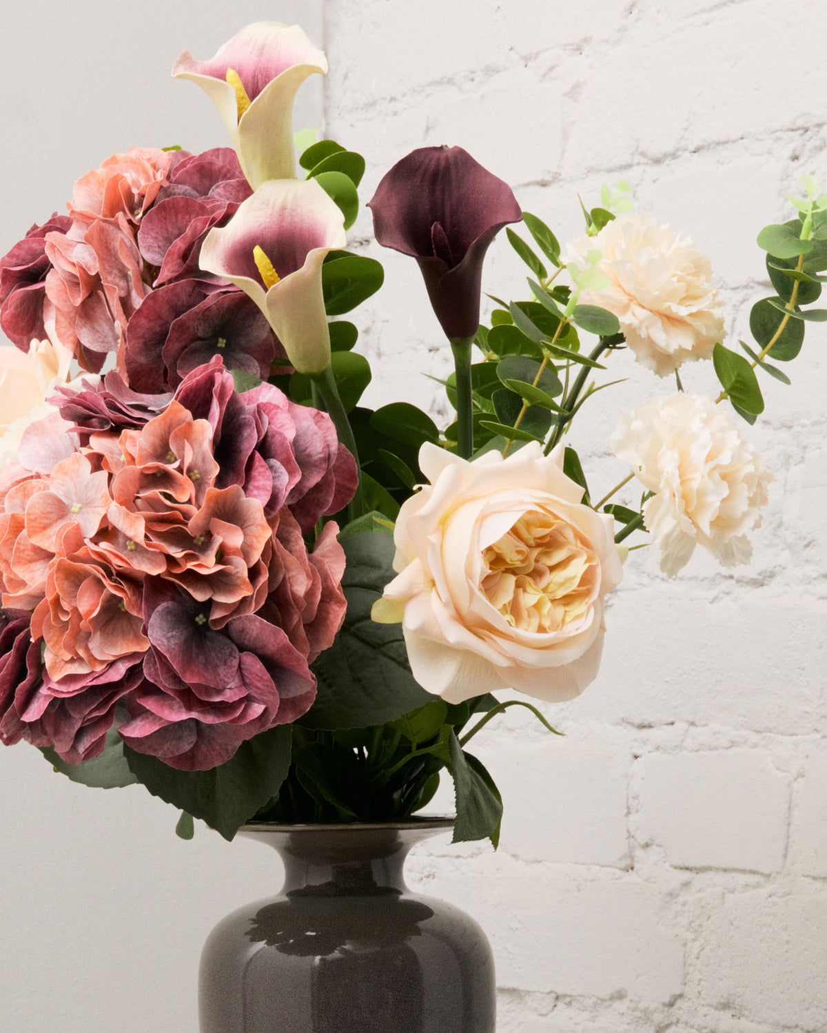 Närbild av Burgundy Blush med konstgjorda antikrosa hortensior, champagnefärgade rosor, vita kallor med lila detaljer och en vinröd kalla.