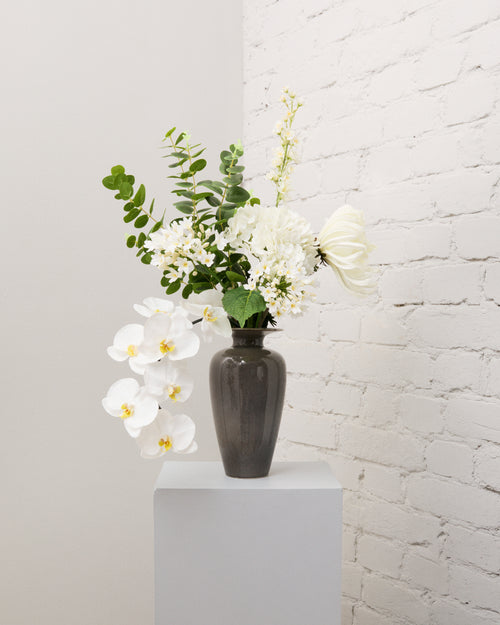 Ivory Grace – konstgjord bukett med vit hortensia, agapanthus, riddarsporre, hängande vit orkidé och grön eukalyptus i grå vas.