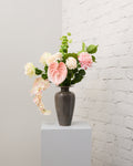 Blush Romance – konstgjord bukett med rosa och krämvita rosor, champagnefärgade nejlikor, rosa anthurium och hängande puderrosa orkidé i grå vas.
