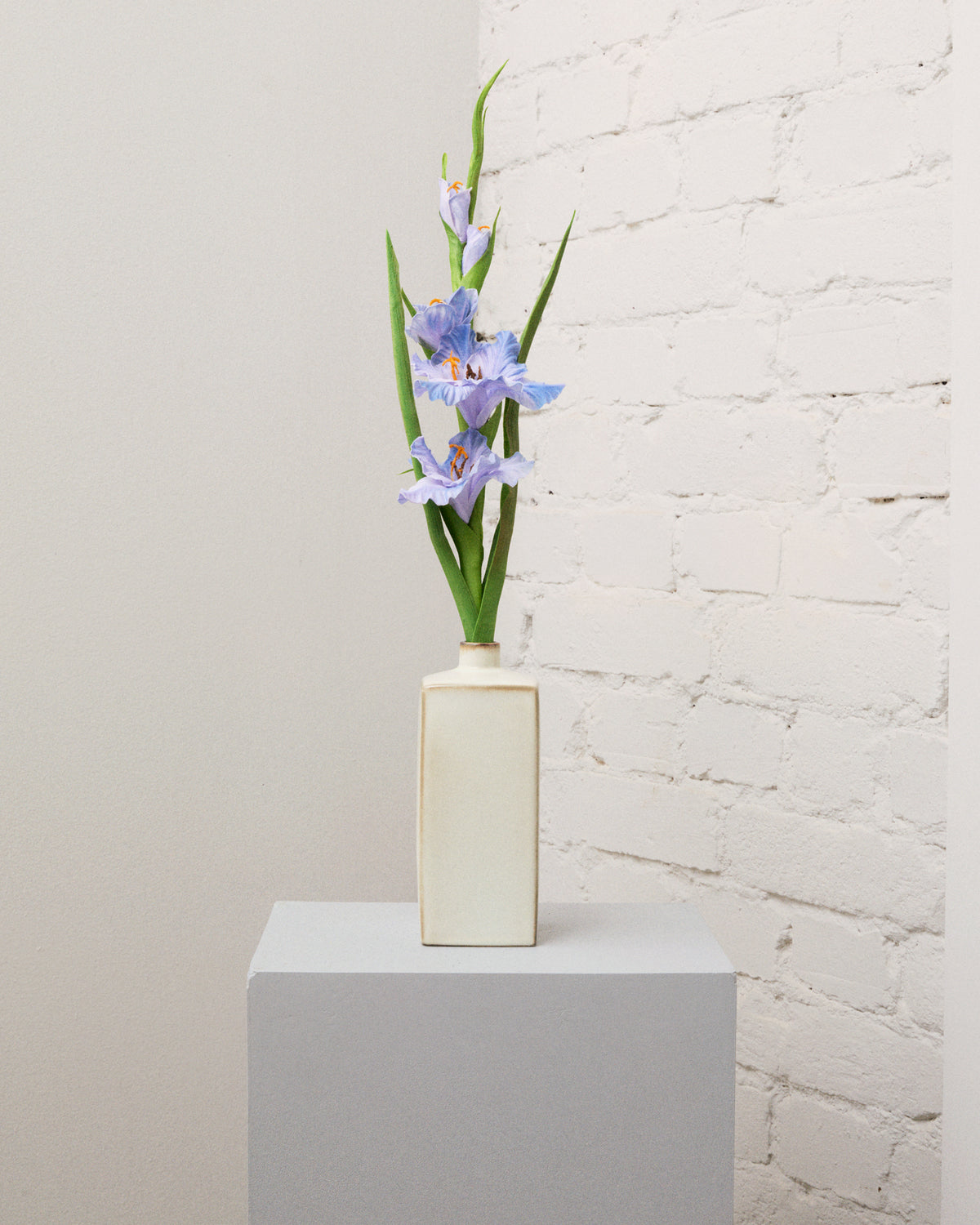 Konstgjord gladiolus i blått med flera blommor, knoppar och långa, smala gröna blad i en krämvit vas.