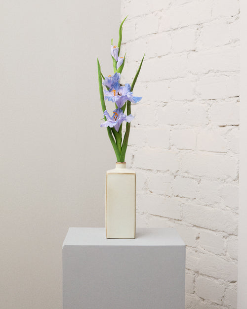 Konstgjord gladiolus i blått med flera blommor, knoppar och långa, smala gröna blad i en krämvit vas.