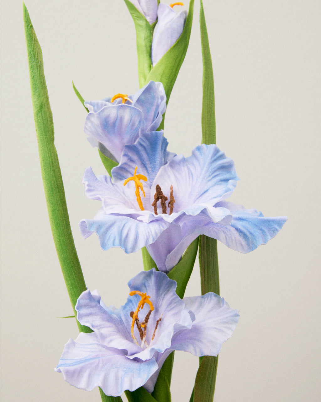 Närbild av konstgjord gladiolus med ljusblå, vågiga kronblad, gula ståndare och långa gröna blad längs stjälken.