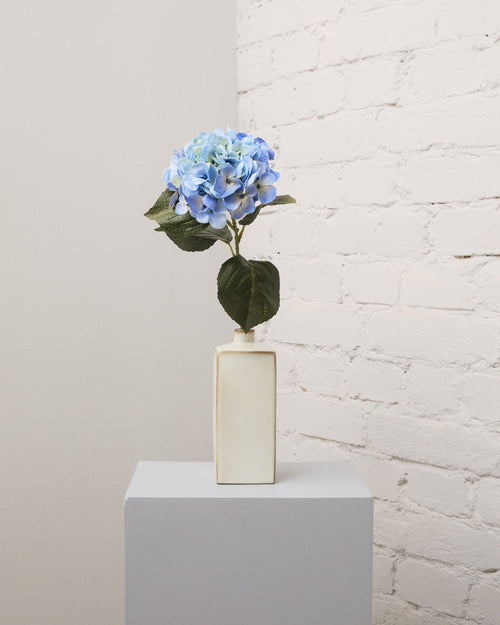 Konstgjord hortensia i blått med ett fylligt, rundat blomhuvud och mörkgröna blad i en smal, krämvit vas.