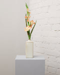 Konstgjord gladiolus i champagnebeige med flera blommor, knoppar och långa, smala gröna blad i en krämvit vas.