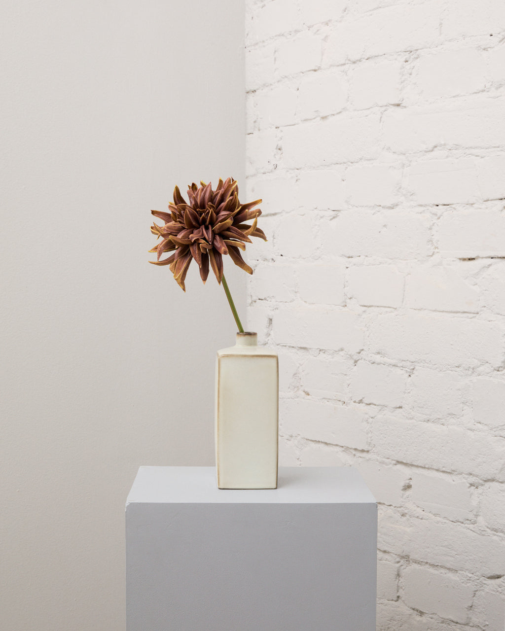 Konstgjord kastanjebrun dahlia med en stor, fyllig blomma på en grön stjälk i en smal, krämvit vas.