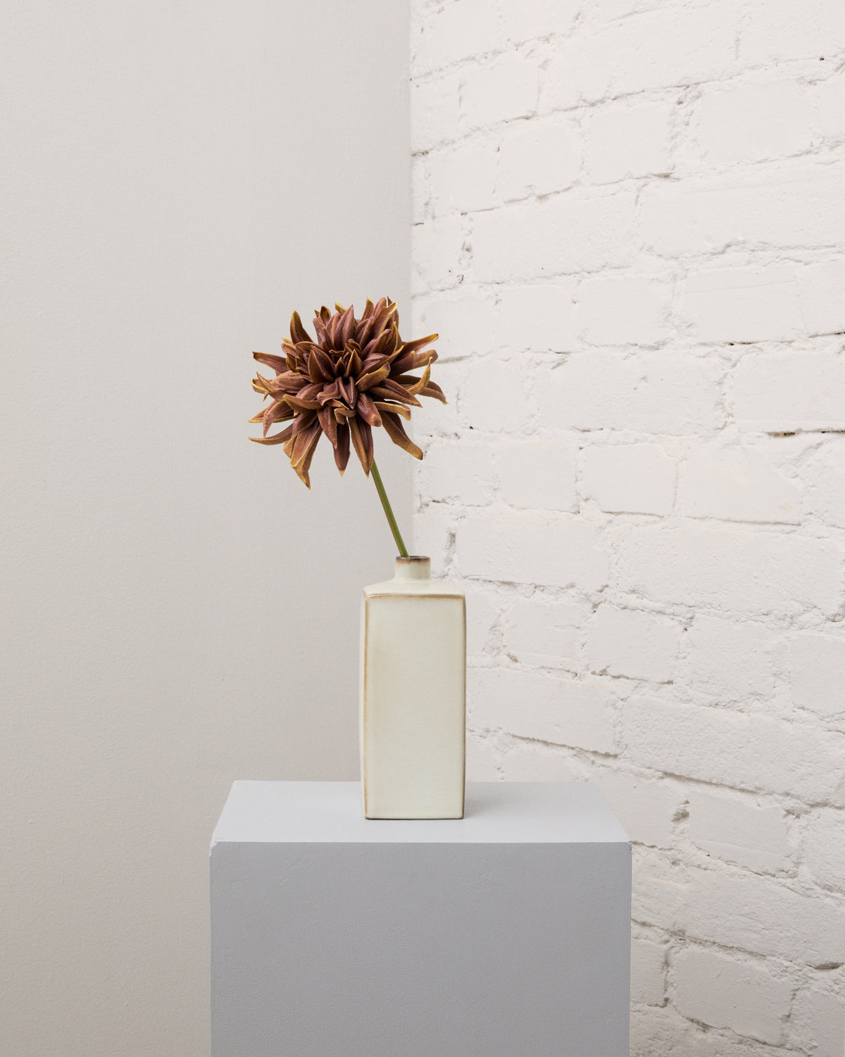 Konstgjord kastanjebrun dahlia med en stor, fyllig blomma på en grön stjälk i en smal, krämvit vas.
