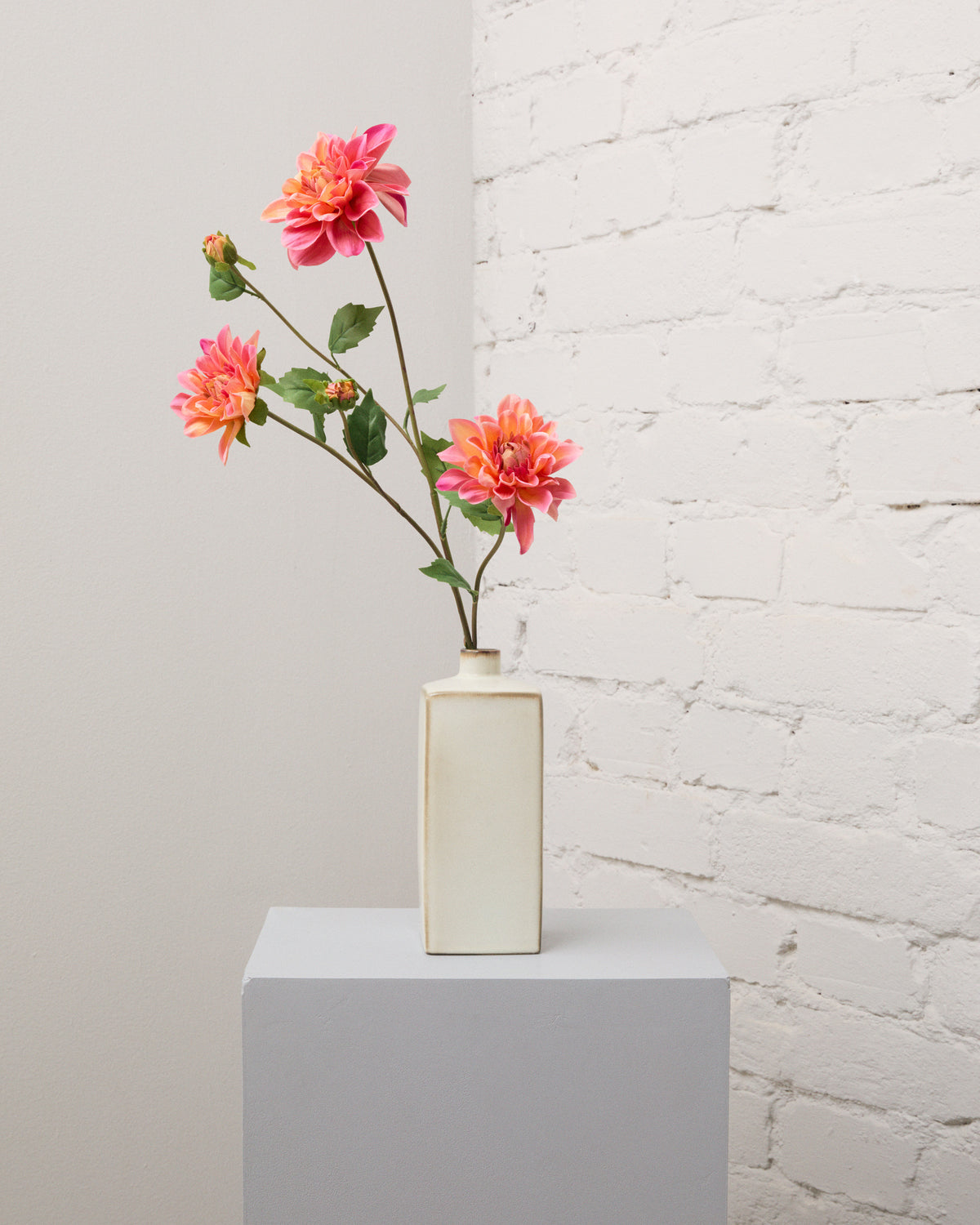 Konstgjord dahlia i orange och korallrosa med flera blommor, knoppar och gröna blad i en smal, krämvit vas.