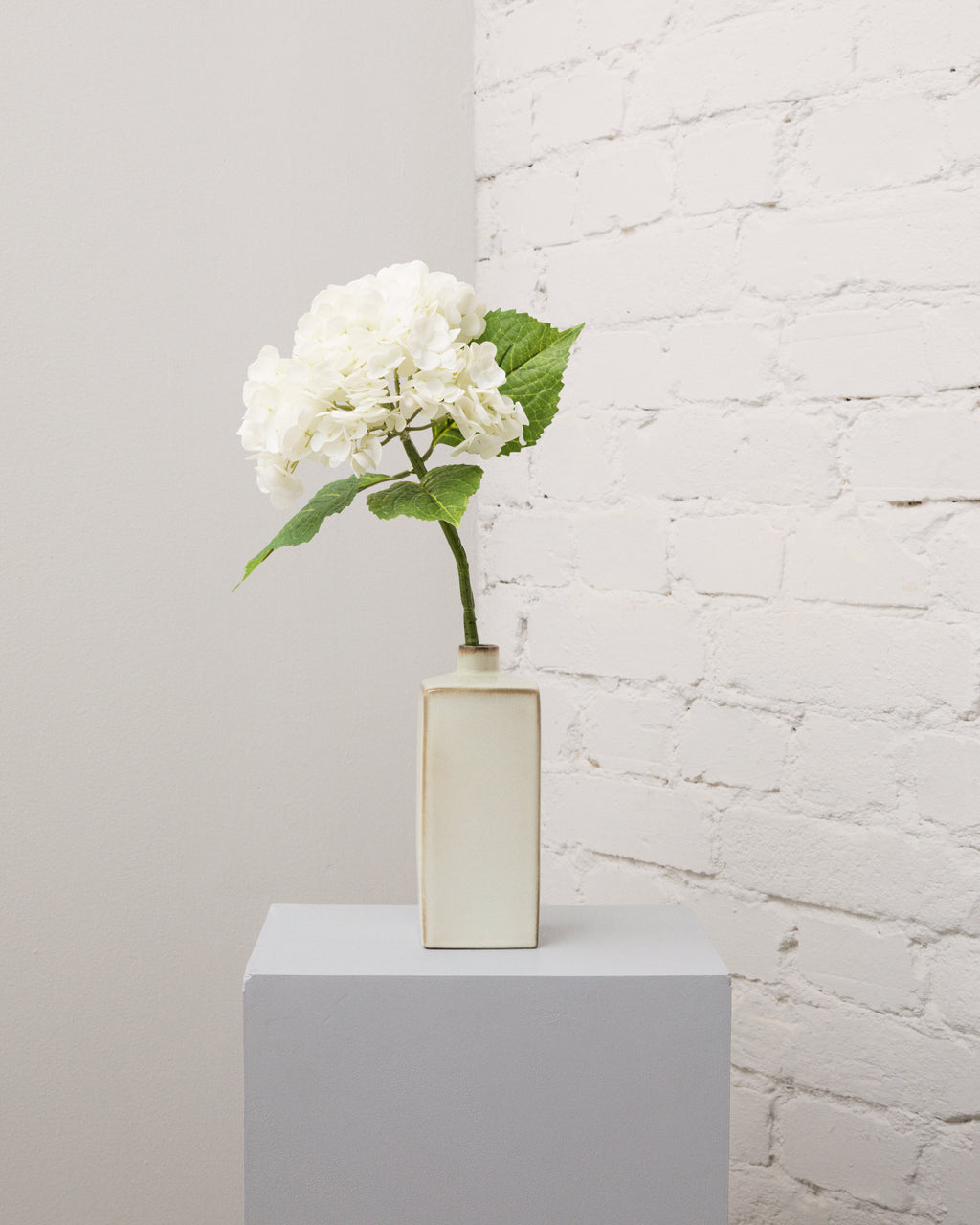 Konstgjord hortensia i vitt med ett stort, fylligt blomhuvud och gröna blad i en smal, krämvit vas.