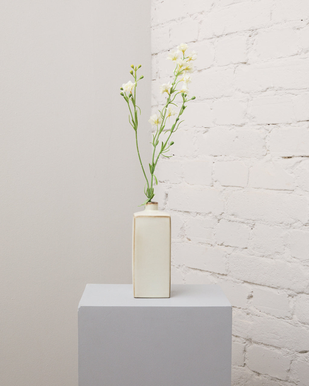 Konstgjord riddarsporre i vitt med små blommor och gröna knoppar på tunna, förgrenade stjälkar i en smal, krämvit vas.