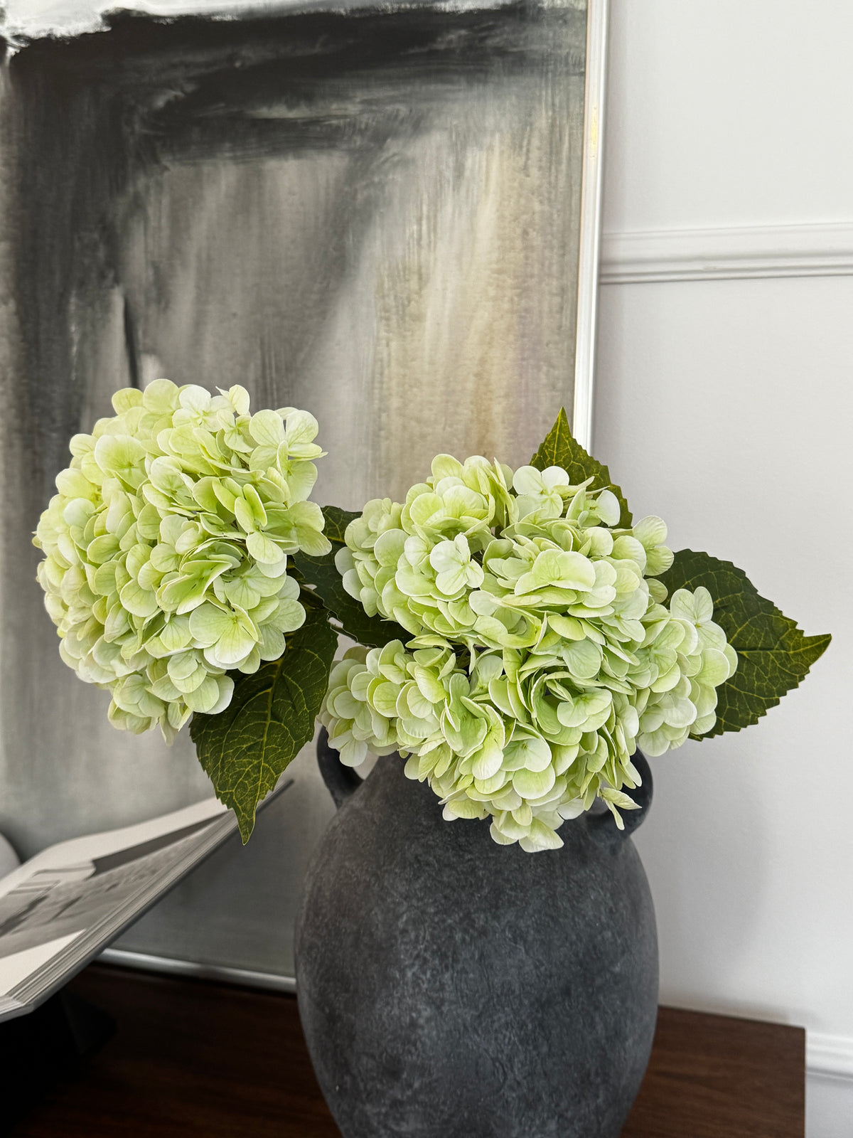 Konstgjord hortensia i ljusgrönt med fylliga blomhuvuden och mörkgröna blad i en mörkgrå vas på ett träbord framför en abstrakt tavla.